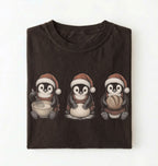 A dark brown folded t-shirt featuring a graphic of three penguins wearing Santa hats, each holding a different baking ingredient (sugar, flour, chocolate), on a clean white background.