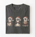 A dark gray folded t-shirt featuring a graphic of three penguins wearing Santa hats and scarves, each holding a bowl or package, on a plain white background.