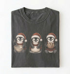 A dark gray folded t-shirt featuring a graphic of three penguins wearing Santa hats and scarves, each holding a bowl or package, on a plain white background.