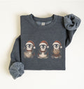 A folded dark heather gray sweatshirt featuring a graphic of three penguins wearing Santa hats, one holding a bowl, one holding a snowball, and one holding a cookie. The sleeves are rolled up and displayed on either side of the sweatshirt against a plain off-white background.