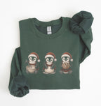 A dark green long-sleeve crewneck sweatshirt featuring a graphic of three penguins wearing Santa hats on the front. The sweatshirt is styled with a matching dark green scrunchie on a clean white background.