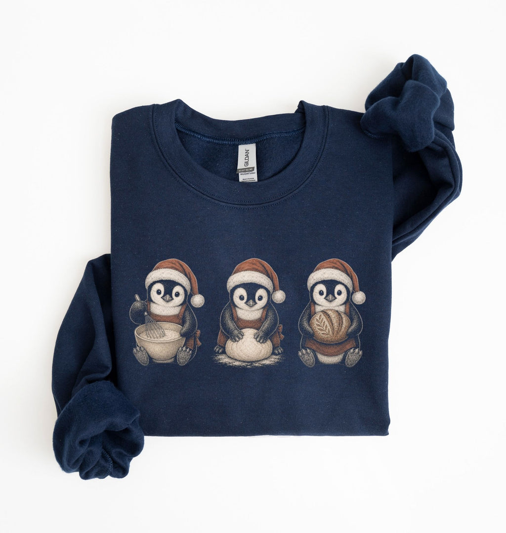 A navy blue long-sleeve sweatshirt with three cartoon penguins wearing Santa hats printed on the chest, shown folded on a white background.