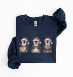 A navy blue long-sleeve sweatshirt with three cartoon penguins wearing Santa hats printed on the chest, shown folded on a white background.