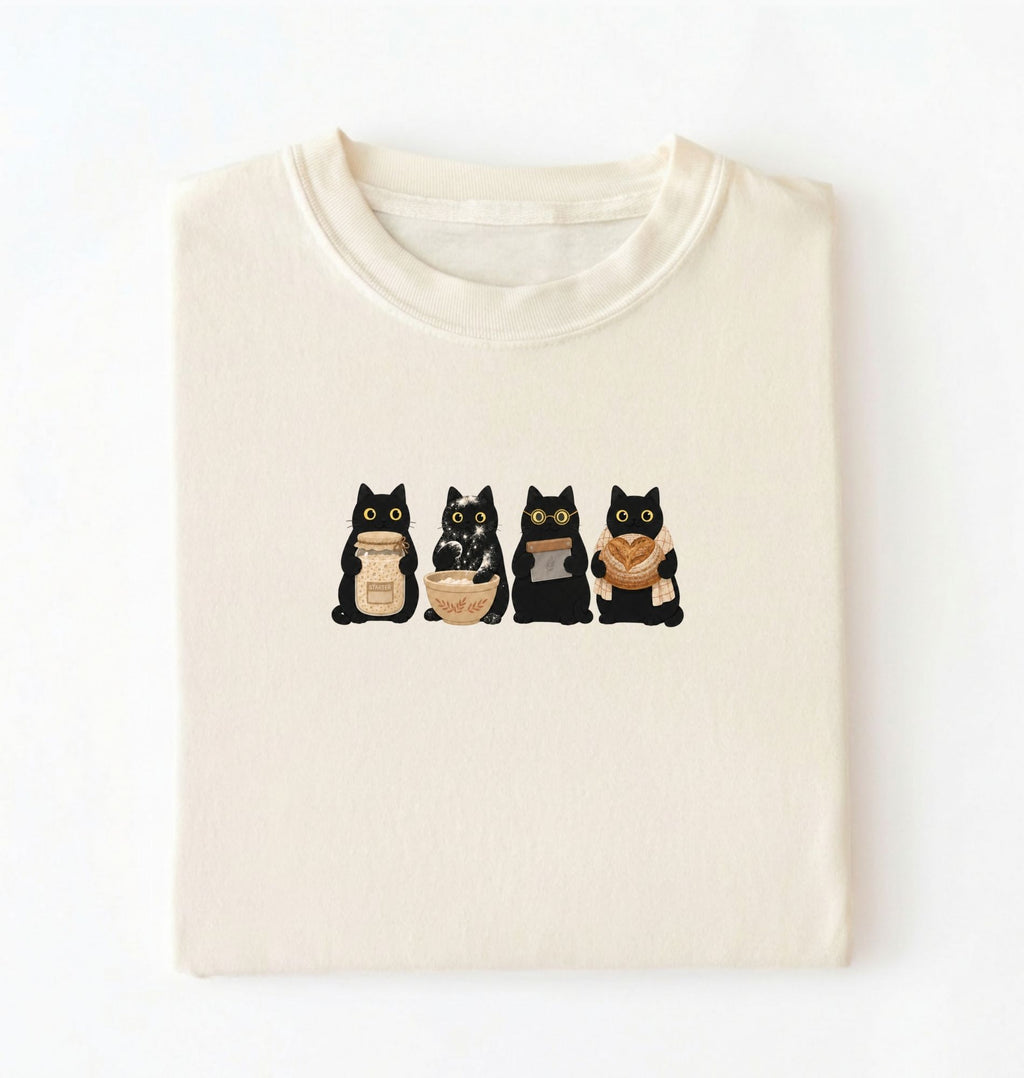 Flat lay of a Cute black cat baker shirt featuring baking cats with bread and mixing bowl on cream t-shirt on a white background 