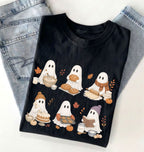 A black short-sleeve T-shirt laid flat on a white surface, styled with light blue jeans at the top. The shirt features a graphic print of six cute cartoon ghosts dressed in autumn clothing, each holding seasonal treats like bread, pumpkin pie, cookies, and a mug, with small orange leaves and pumpkins scattered around.