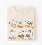 A cream-colored crewneck t-shirt with a graphic print of five cute cartoon ghosts baking autumn treats, displayed flat against a clean white background.