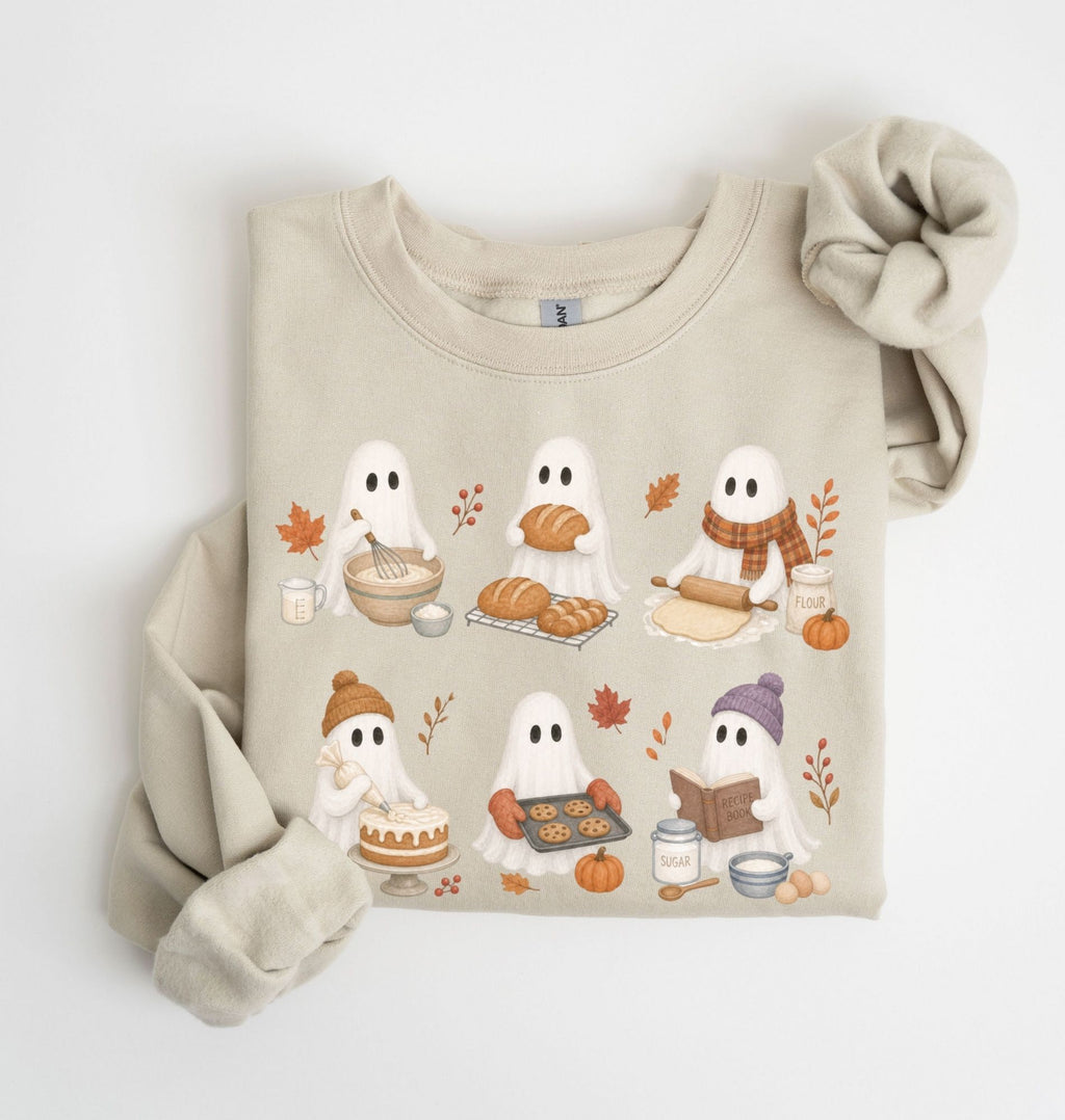A beige crewneck sweatshirt featuring a cute cartoon graphic of five ghosts baking and playing with autumn elements, styled flat on a white background with a matching beige knit scrunchie.