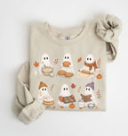 A beige crewneck sweatshirt featuring a cute cartoon graphic of five ghosts baking and playing with autumn elements, styled flat on a white background with a matching beige knit scrunchie.