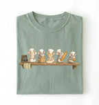 A folded sage green t-shirt with a graphic print of five cute cartoon mice dressed as chefs and bakers sitting on a wooden shelf, displayed against a plain white background.