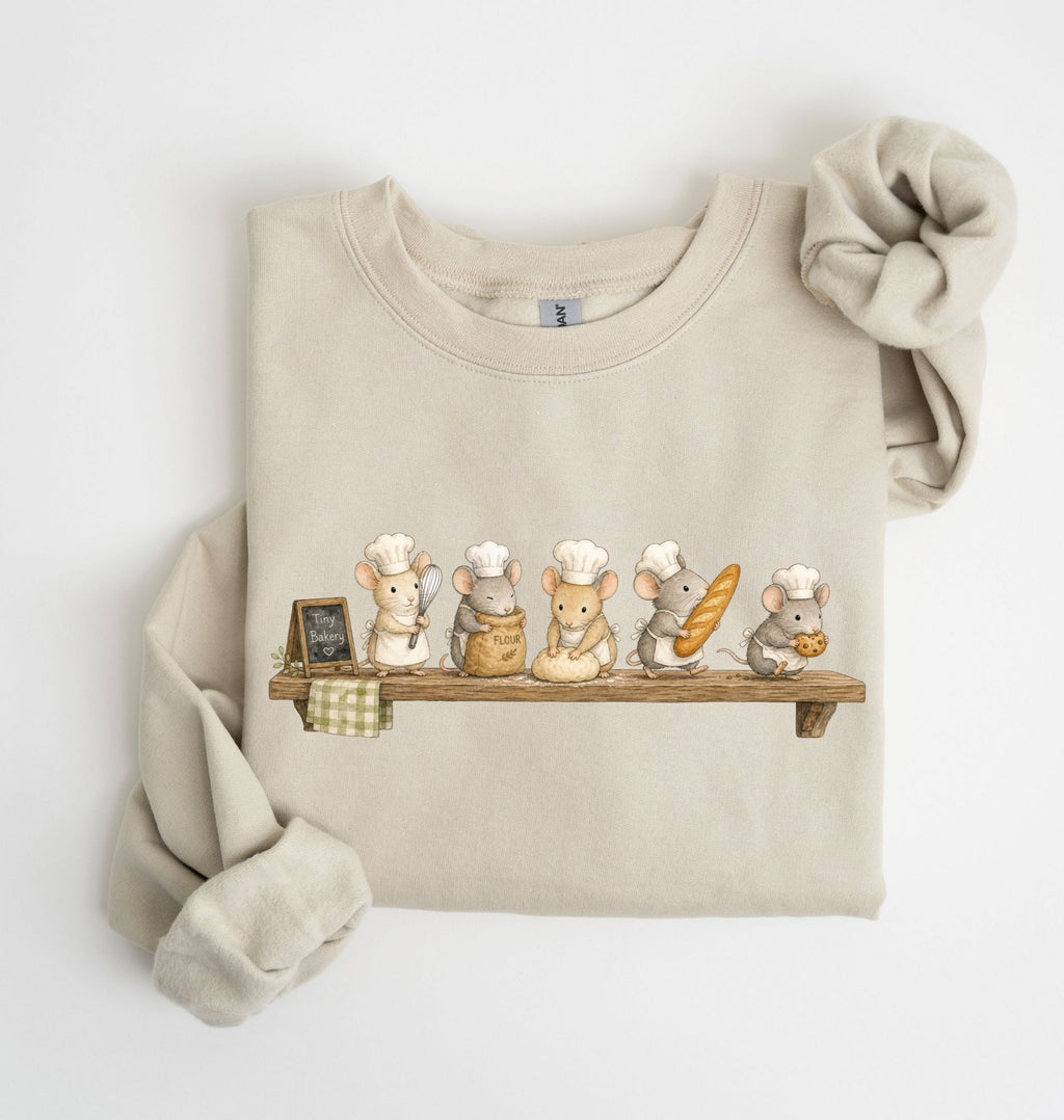 A beige crewneck sweatshirt with a graphic of five cute cartoon mice dressed as chefs sitting on a wooden shelf, styled with a matching beige knit scrunchie on a white background.