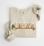 A beige crewneck sweatshirt with a graphic of five cute cartoon mice dressed as chefs sitting on a wooden shelf, styled with a matching beige knit scrunchie on a white background.