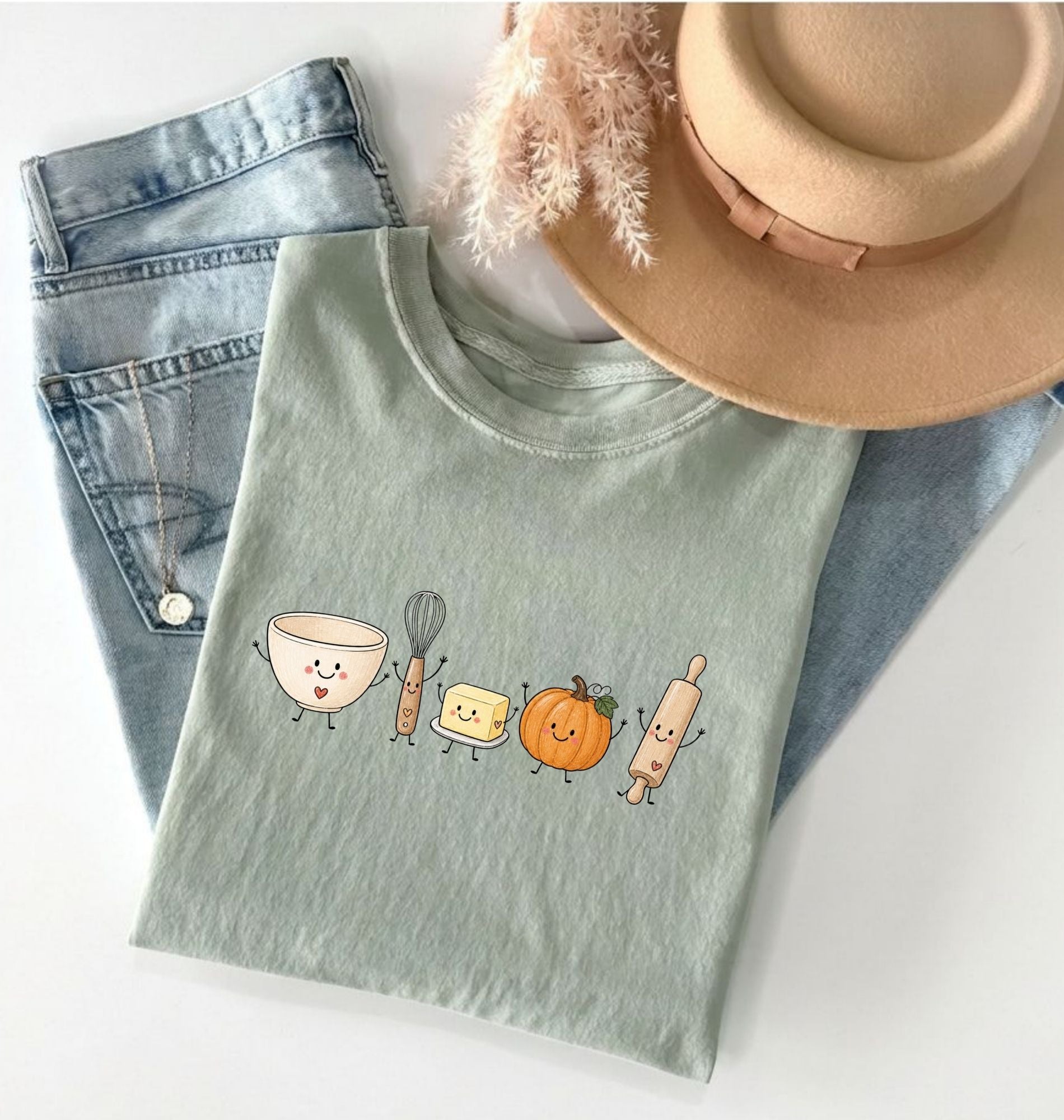 A folded sage green t-shirt with a cute cartoon kitchen utensil graphic, styled in a flat lay with light-wash jeans, a beige hat, and a necklace on a white background.