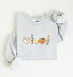 A light heather gray sweatshirt with long sleeves, folded neatly on a white background. The front of the sweatshirt features a cute graphic of a bowl, whisk, butter, pumpkin, and rolling pin with smiling faces.