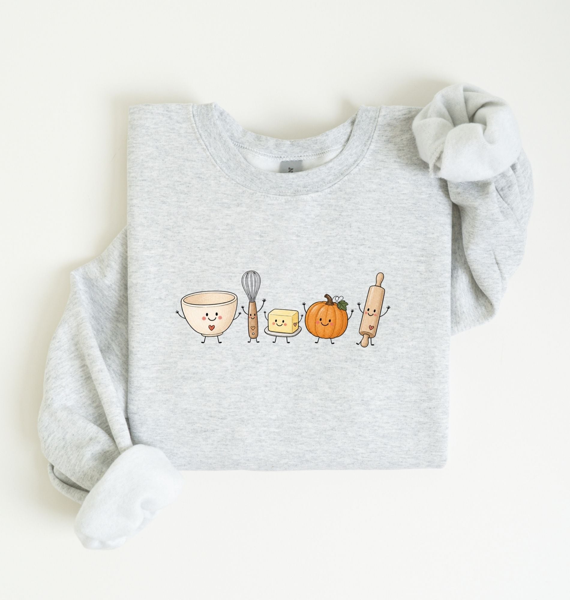 A light heather gray sweatshirt with long sleeves, folded neatly on a white background. The front of the sweatshirt features a cute graphic of a bowl, whisk, butter, pumpkin, and rolling pin with smiling faces.