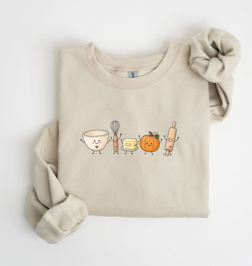 A folded beige crewneck sweatshirt featuring a cute cartoon graphic of baking ingredients (a bowl, whisk, butter, pumpkin, and rolling pin) with faces, styled with a matching beige scrunchie on a white background.