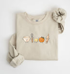 A folded beige crewneck sweatshirt featuring a cute cartoon graphic of baking ingredients (a bowl, whisk, butter, pumpkin, and rolling pin) with faces, styled with a matching beige scrunchie on a white background.