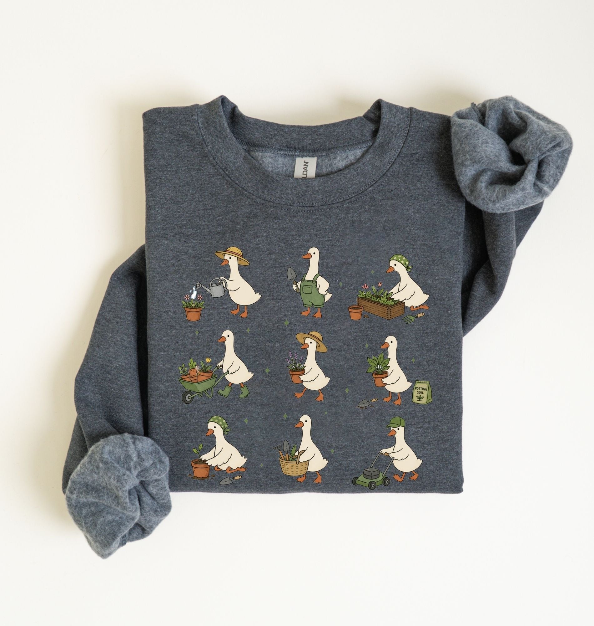 A folded dark heather gray sweatshirt featuring a graphic print of white garden geese with orange beaks and feet, gardening with pots, tools, and carts. The sleeves are styled rolled up, and the sweatshirt is laid flat on a clean off-white background.