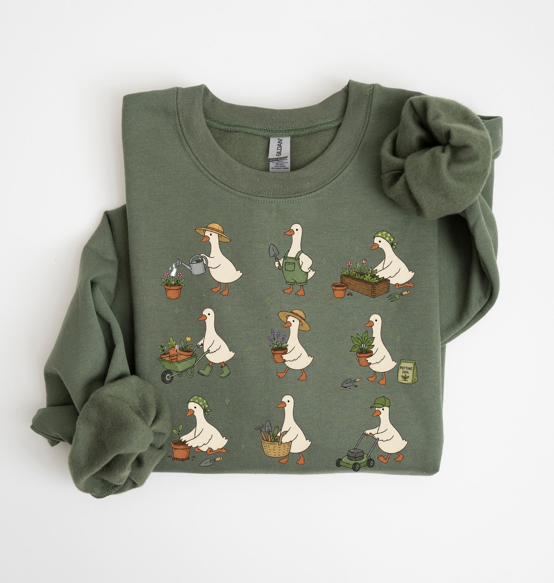 An olive green crewneck sweatshirt featuring a grid pattern of cartoon garden geese, folded neatly on a white background with a matching fabric flower accessory tucked into one sleeve.
