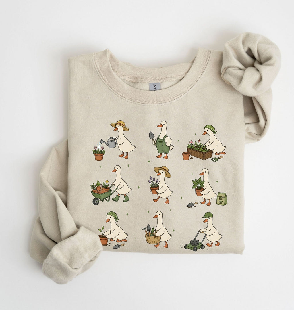 A folded beige crewneck sweatshirt featuring a cute cartoon pattern of white geese doing gardening activities like watering plants, pushing a wheelbarrow, and mowing grass. The sweatshirt is styled with a matching beige knit scrunchie on a clean white background.