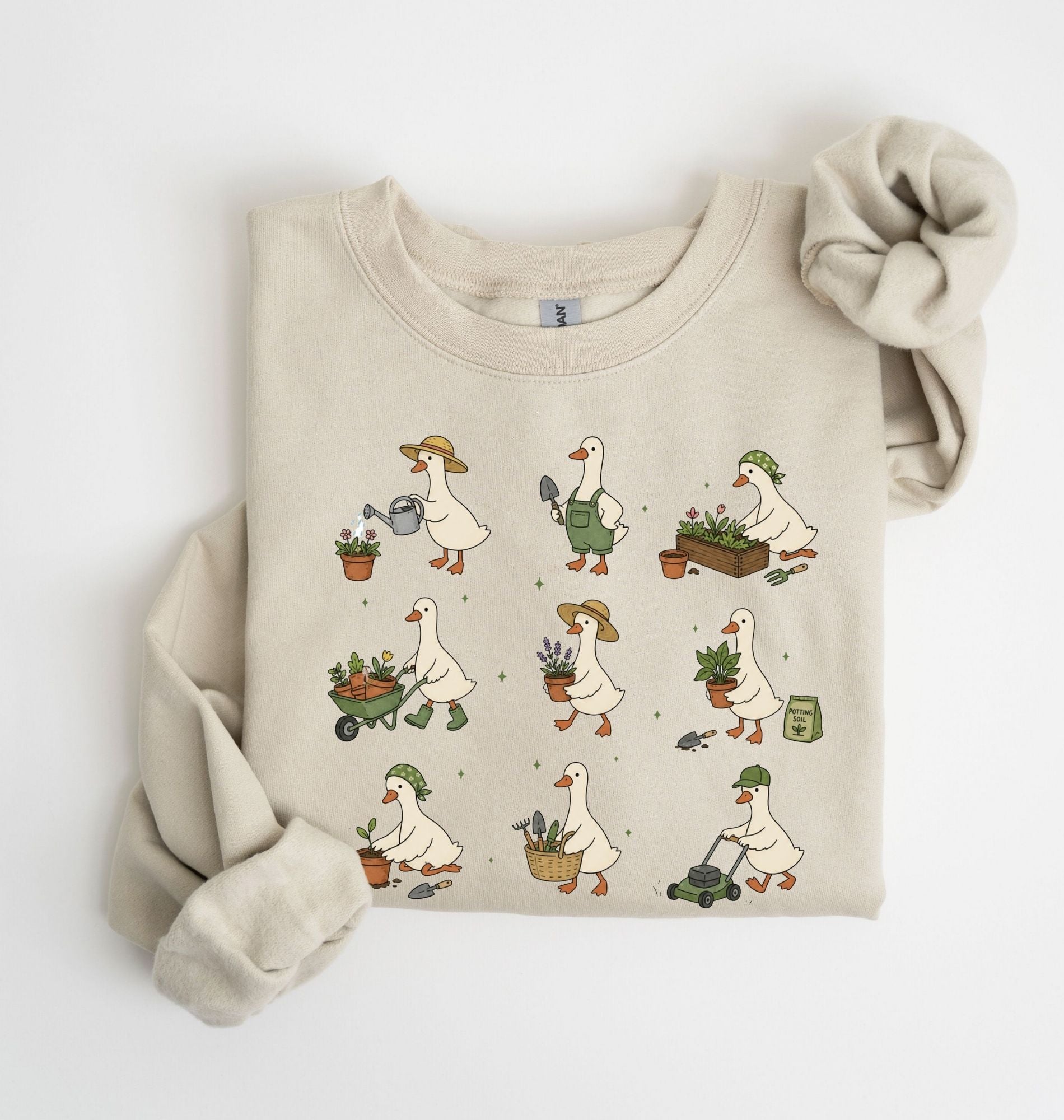 A folded beige crewneck sweatshirt featuring a cute cartoon pattern of white geese doing gardening activities like watering plants, pushing a wheelbarrow, and mowing grass. The sweatshirt is styled with a matching beige knit scrunchie on a clean white background.