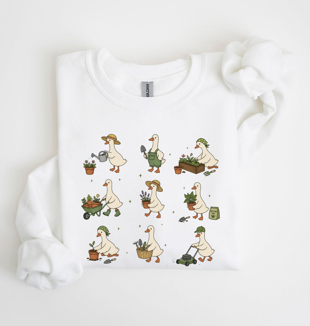 A white long-sleeve crewneck sweatshirt with a graphic print of nine cartoon geese doing gardening activities, laid flat on a white background with a matching white fabric prop.