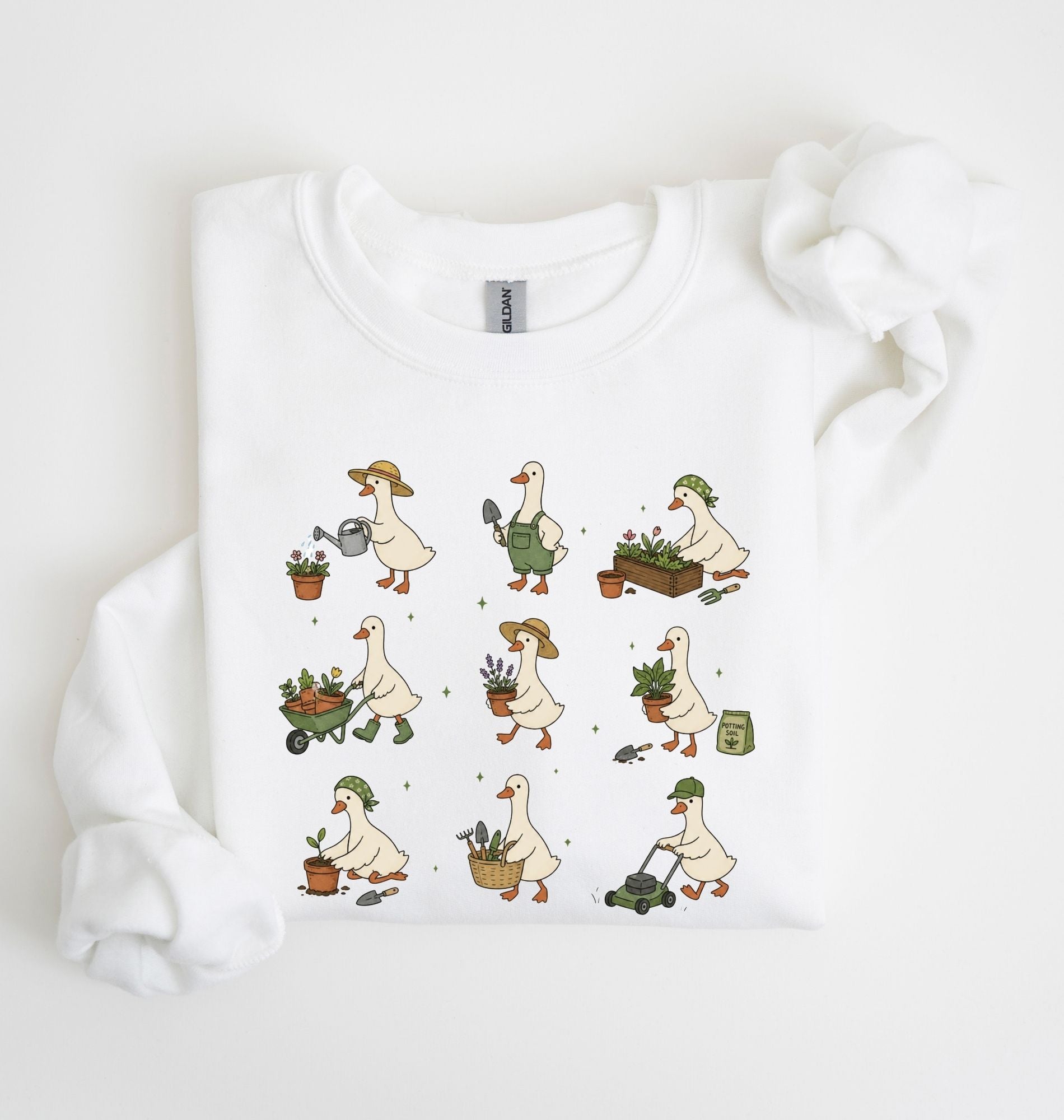 A white long-sleeve crewneck sweatshirt with a graphic print of nine cartoon geese doing gardening activities, laid flat on a white background with a matching white fabric prop.