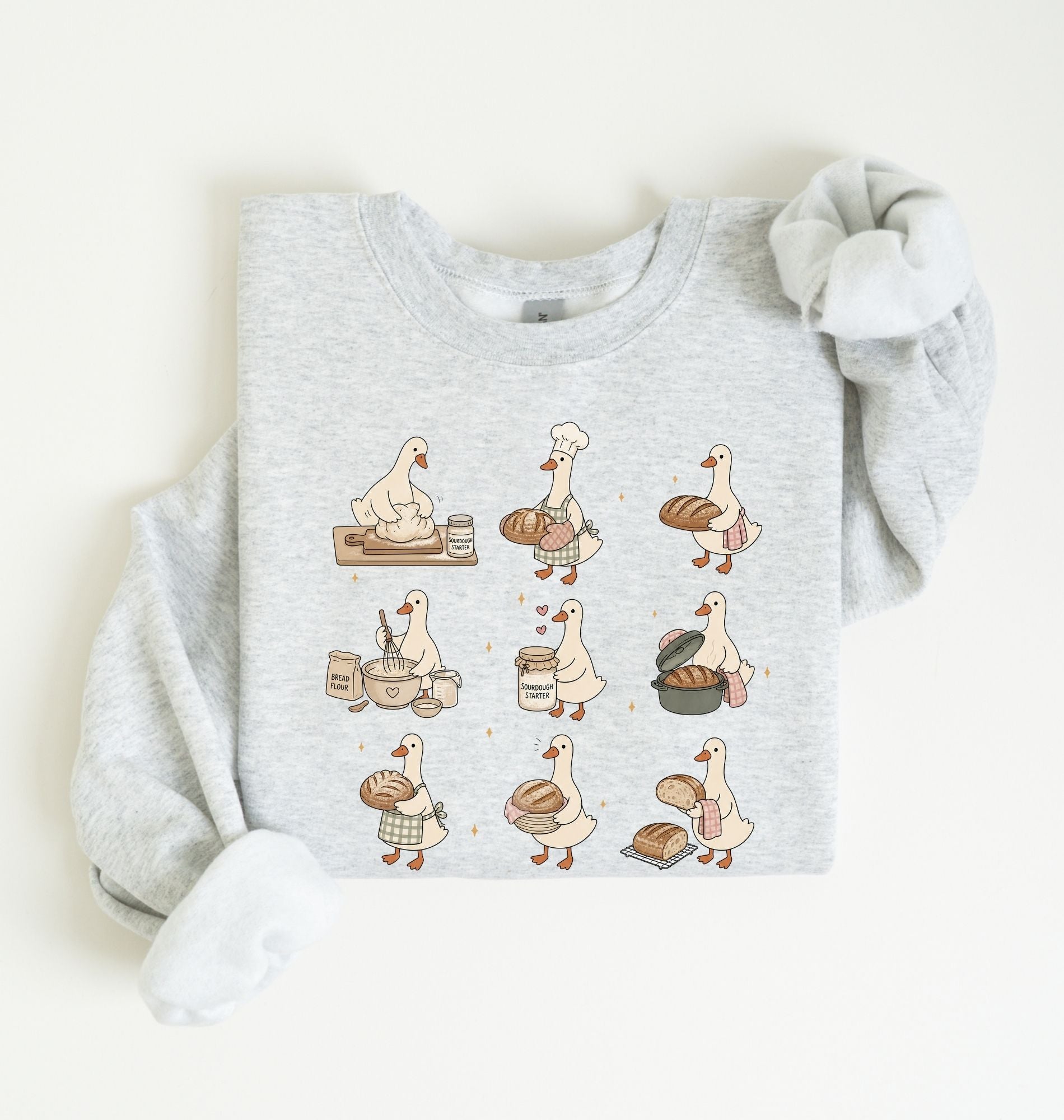 A light heather gray long-sleeve sweatshirt with a graphic print of nine cartoon geese in various baking and bread-related poses, styled flat on a light cream background.