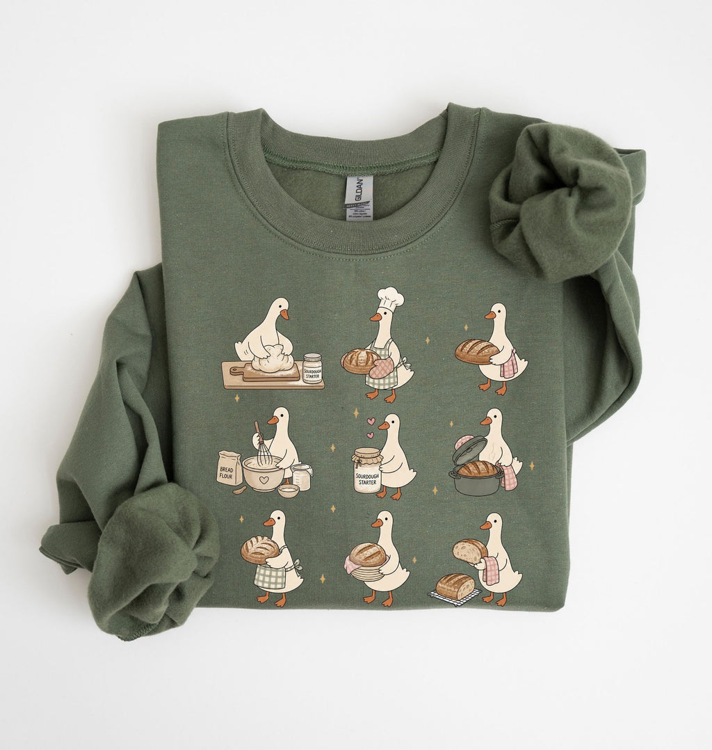 An olive green long-sleeve sweatshirt with a graphic print of nine bakers' ducks in various cooking and baking poses, shown folded on a white background.