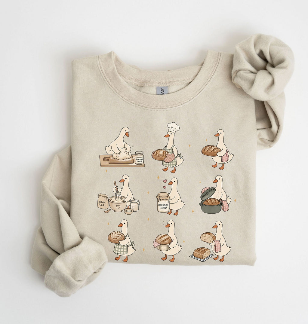 A beige crewneck sweatshirt featuring a cartoon graphic of a goose baking bread in a nine-step sequence, styled flat on a white background with a matching beige knit scrunchie.