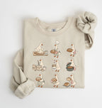 A beige crewneck sweatshirt featuring a cartoon graphic of a goose baking bread in a nine-step sequence, styled flat on a white background with a matching beige knit scrunchie.