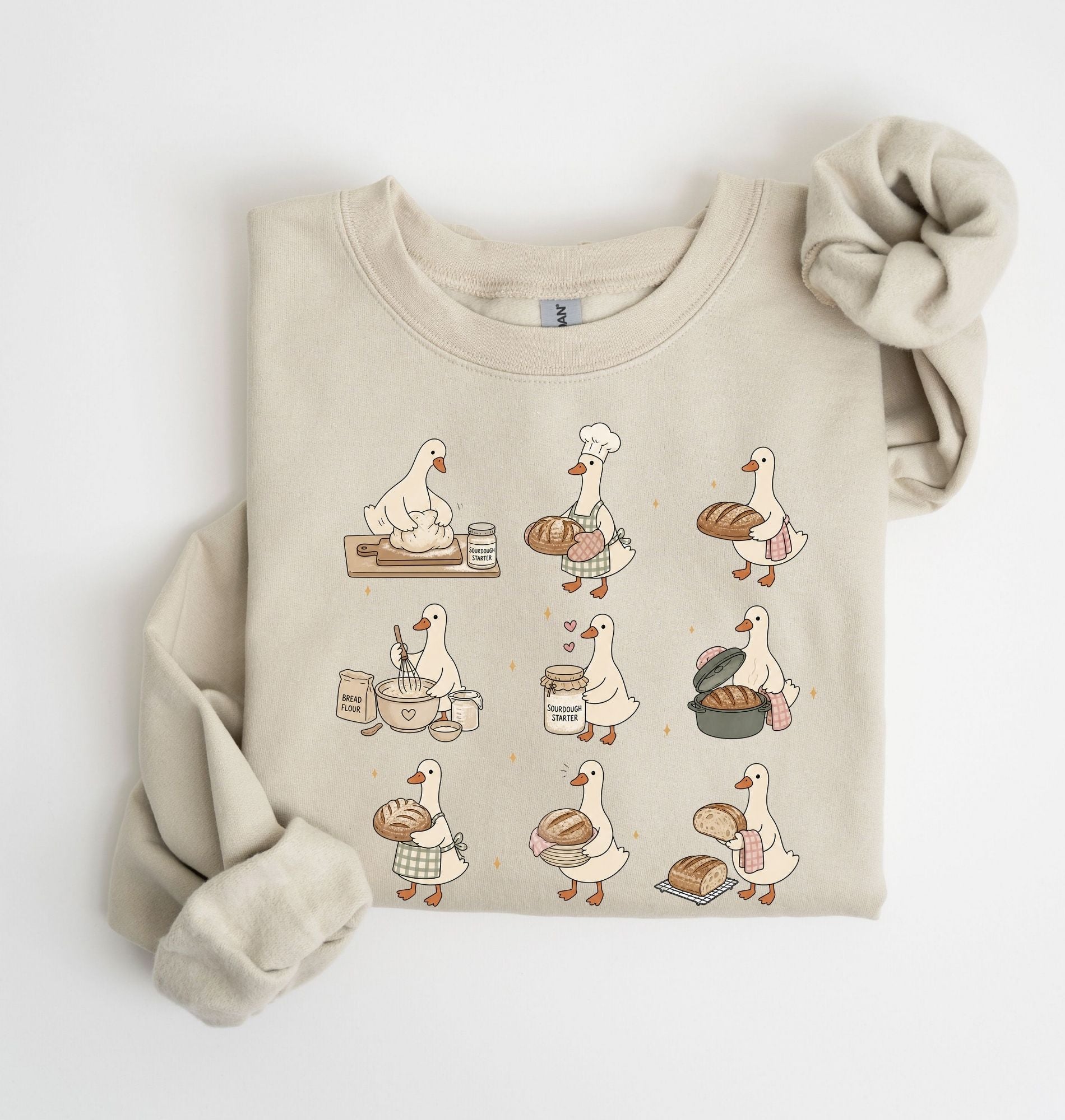 A beige crewneck sweatshirt featuring a cartoon graphic of a goose baking bread in a nine-step sequence, styled flat on a white background with a matching beige knit scrunchie.