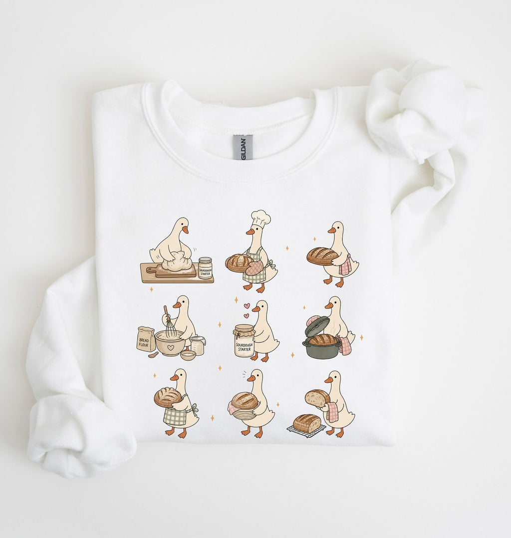 A white crewneck sweatshirt with a graphic print of nine cartoon ducks dressed as bakers, shown folded on a white background with a matching white ribbon.