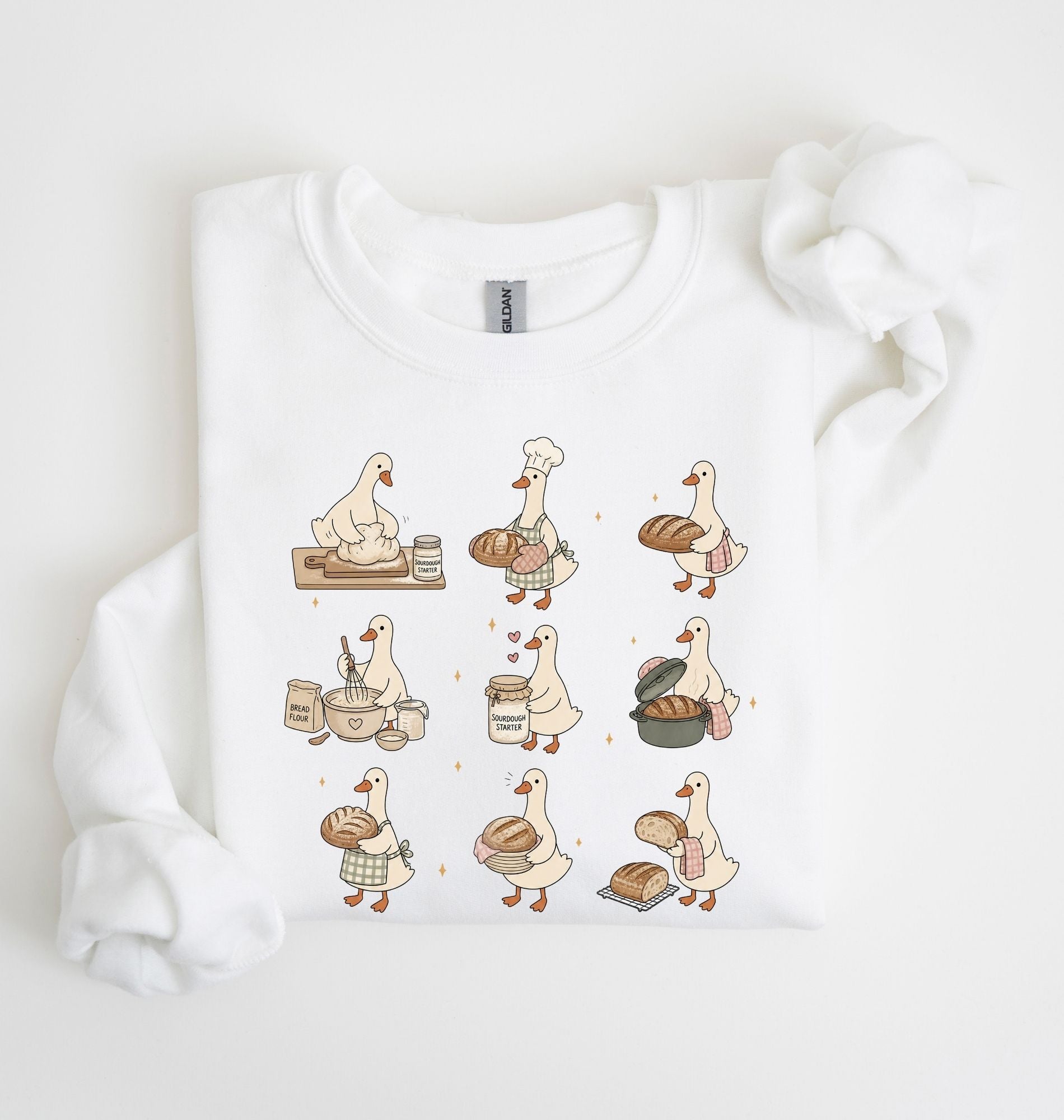 A white crewneck sweatshirt with a graphic print of nine cartoon ducks dressed as bakers, shown folded on a white background with a matching white ribbon.