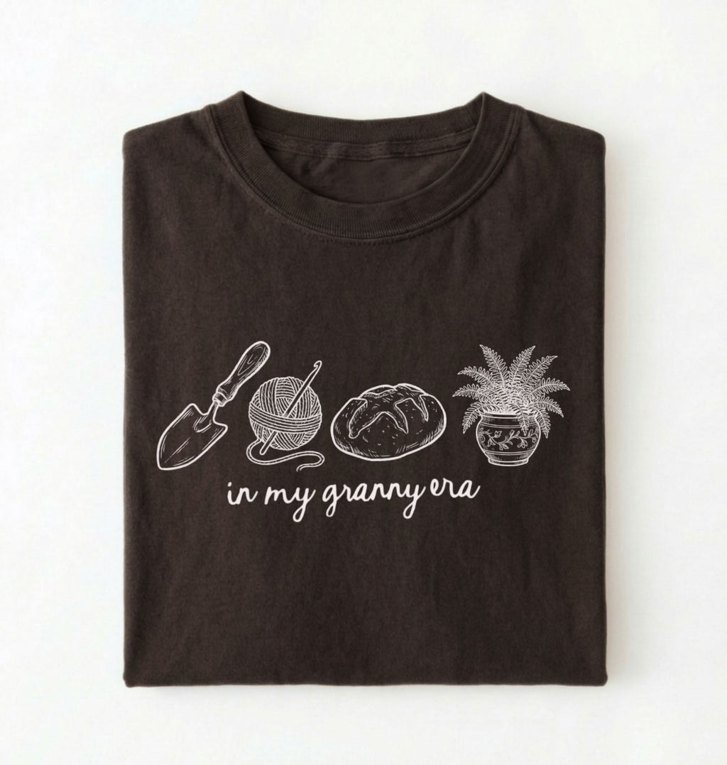 Flat lay of a dark chocolate t-shirt featuring delicate line drawings of gardening tools, yarn, bread, and a plant pot with text reading “in my granny era.”