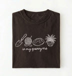 Flat lay of a dark chocolate t-shirt featuring delicate line drawings of gardening tools, yarn, bread, and a plant pot with text reading “in my granny era.”