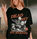A woman wearing a black oversized graphic t-shirt with a distressed orange and white print that reads 'DEAD SET ON BAKING' featuring a skeleton baking illustration. She is posing in an elevator next to the metal button panel.