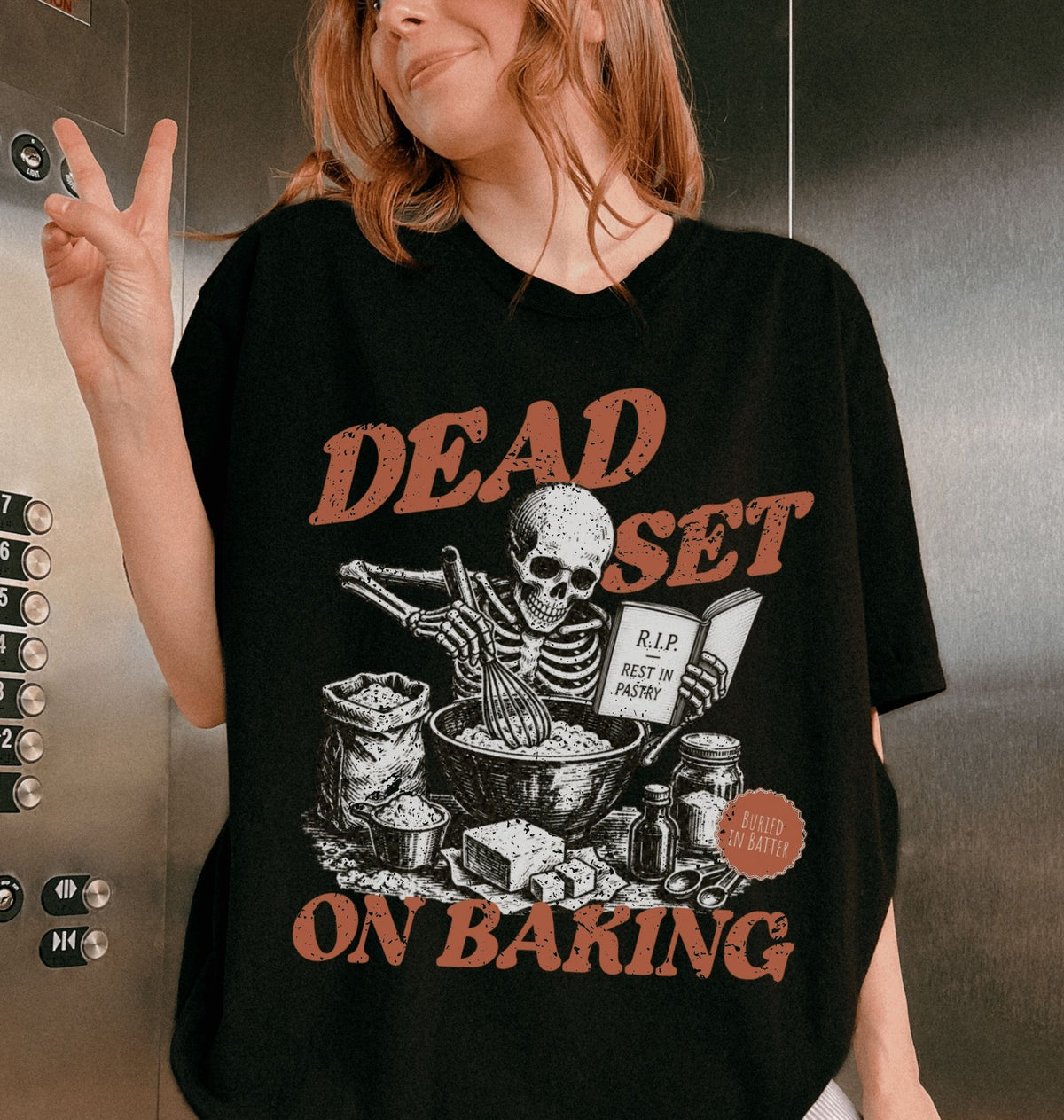 A woman wearing a black oversized graphic t-shirt with a distressed orange and white print that reads 'DEAD SET ON BAKING' featuring a skeleton baking illustration. She is posing in an elevator next to the metal button panel.