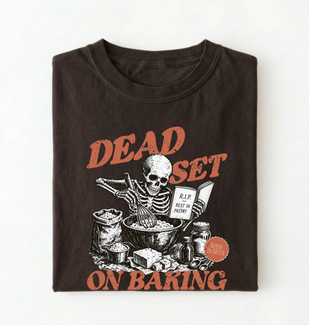 A folded dark brown t-shirt with a graphic print of a skeleton baking, featuring the text 'DEAD SET ON BAKING' in a distressed orange font.