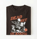 A folded dark brown t-shirt with a graphic print of a skeleton baking, featuring the text 'DEAD SET ON BAKING' in a distressed orange font.