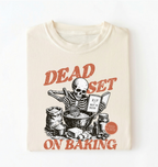 A cream-colored t-shirt folded flat, featuring a graphic print of a skeleton baking with the text 'DEAD SET ON BAKING' in a distressed reddish-brown font.