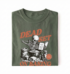 A folded dark olive green t-shirt with a graphic print of a skeleton baking, featuring orange text that reads 'DEAD SET ON BAKING' and a small orange circular badge that says 'KID IN BAKERY'.