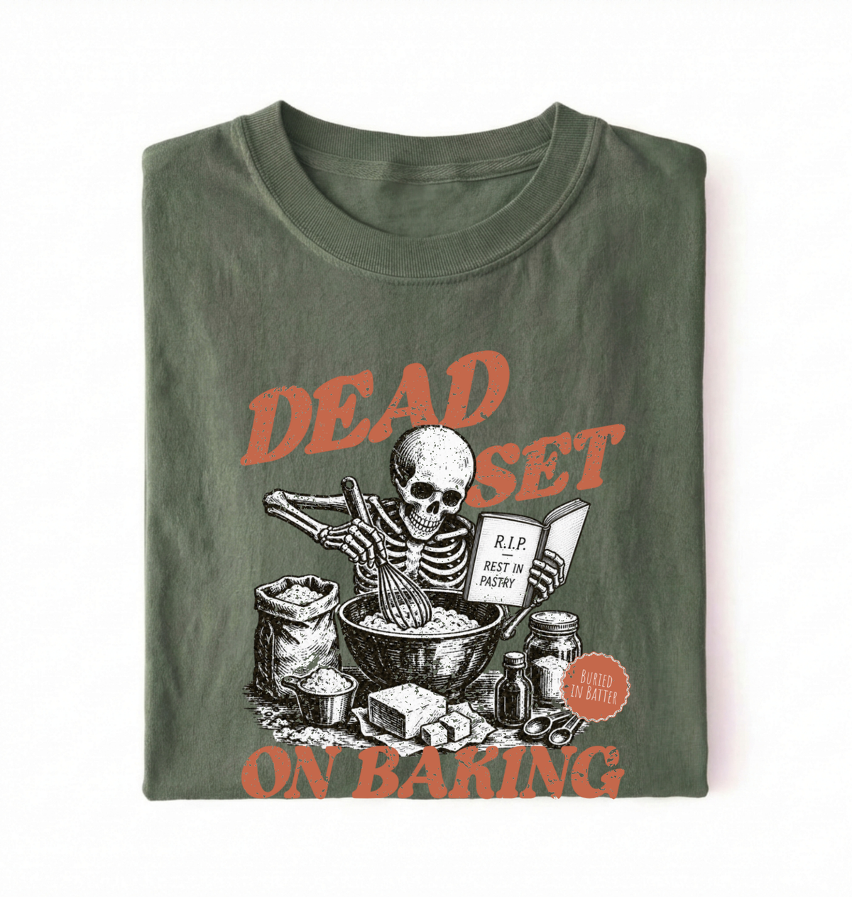 A folded dark olive green t-shirt with a graphic print of a skeleton baking, featuring orange text that reads 'DEAD SET ON BAKING' and a small orange circular badge that says 'KID IN BAKERY'.