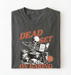 A folded dark gray t-shirt with a graphic print of a skeleton baking, featuring orange text that reads 'DEAD SET ON BAKING' and a small circular badge that says 'MADE IN MASS'.