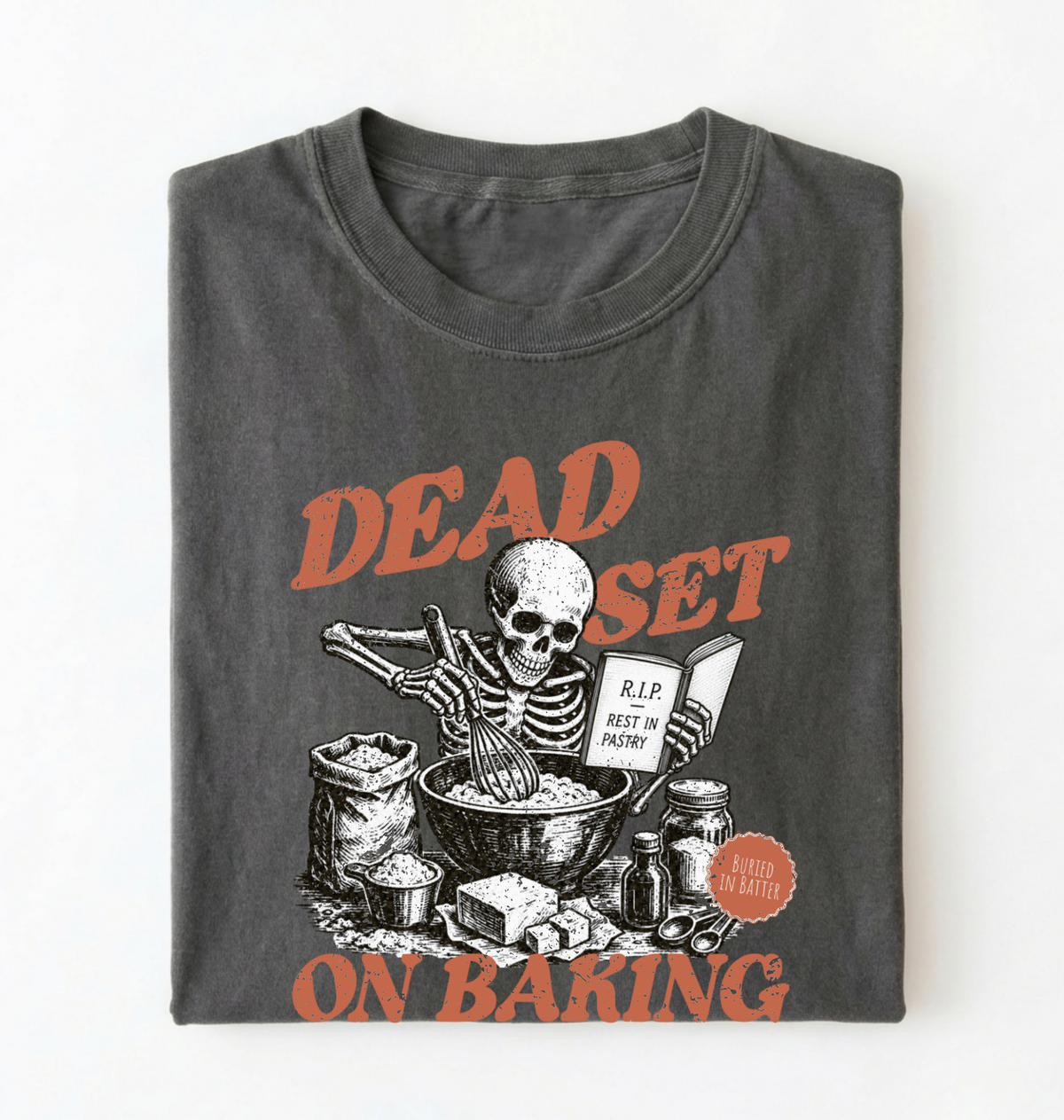 A folded dark gray t-shirt with a graphic print of a skeleton baking, featuring orange text that reads 'DEAD SET ON BAKING' and a small circular badge that says 'MADE IN MASS'.