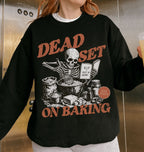 A person wearing a black crewneck sweatshirt with a distressed orange and white graphic that reads 'DEAD SET ON BAKING' around a skeleton baking illustration. The person is holding a drink in a metallic elevator lobby.