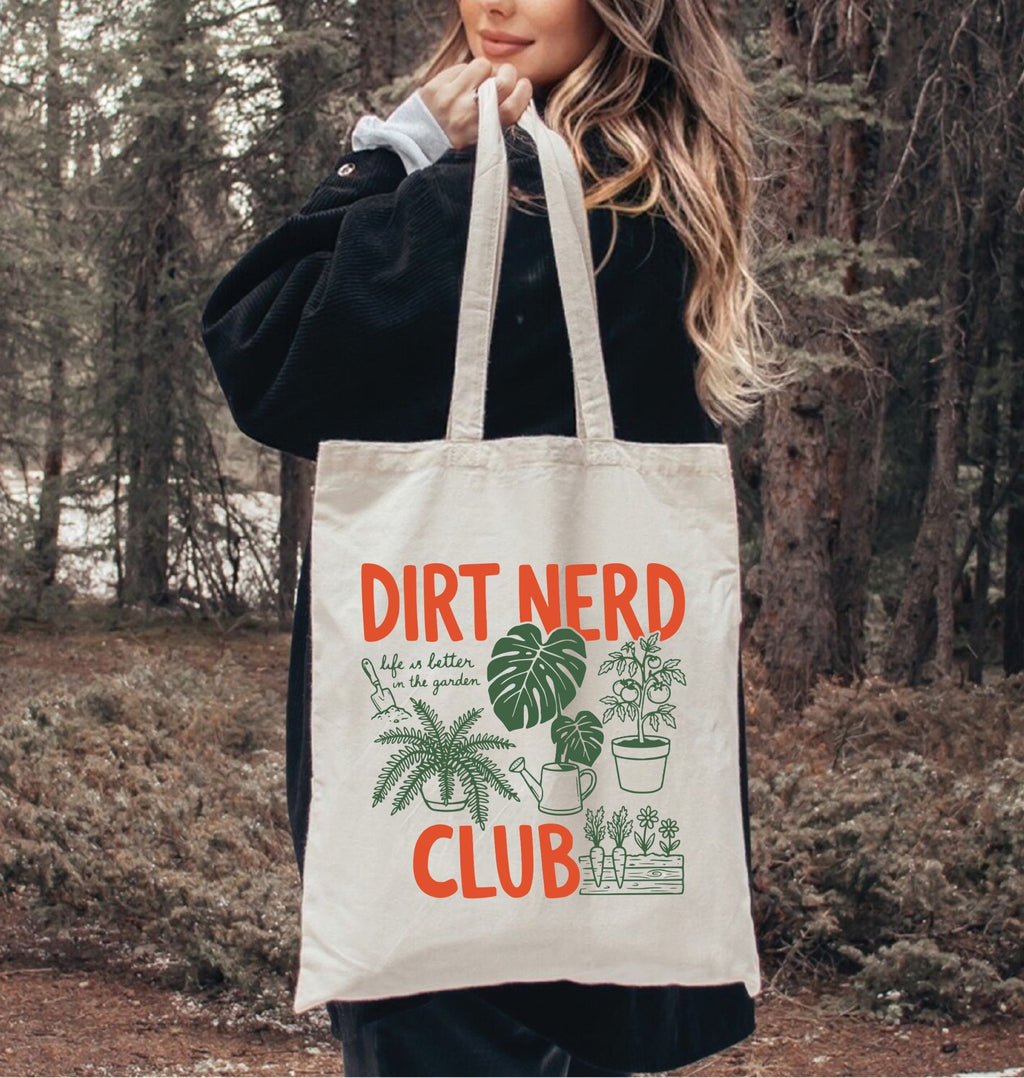 Person holding a tote bag with 'Dirt Nerd Club' text and plant illustrations in a forest setting