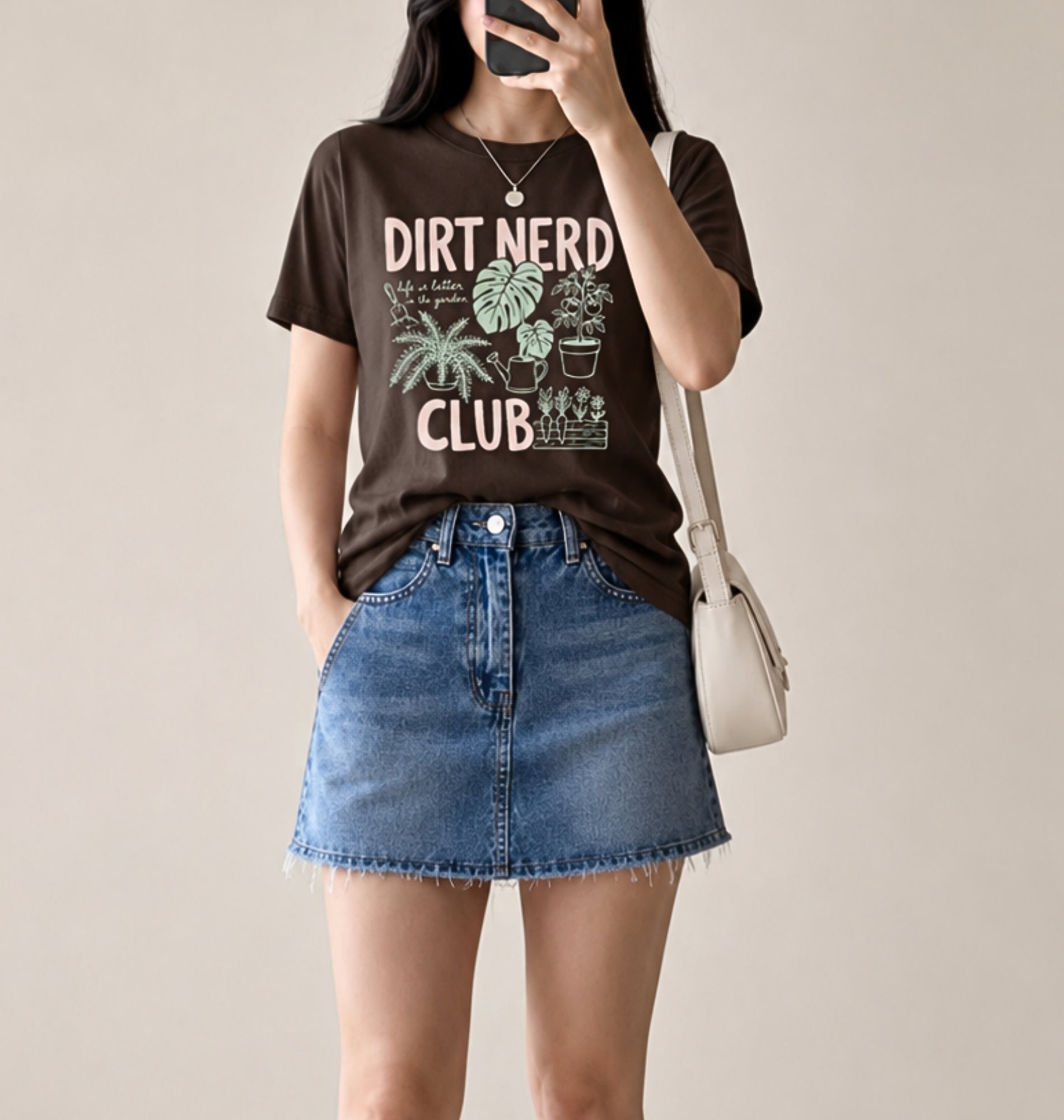 A woman wearing a dark brown graphic t-shirt with 'DIRT NERD CLUB' and plant illustrations, paired with a blue denim mini skirt and a white shoulder bag, shown in a mirror selfie style against a plain beige background.