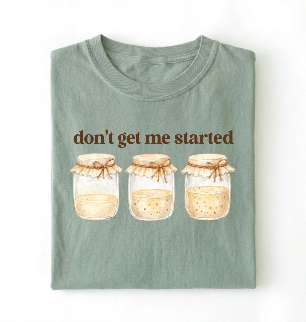 Flat lay of a bay t-shirt featuring illustrated jars and text reading “Don’t Get Me Started. Funny sourdough starter perfect gift for bakers fermentation lovers and bread enthusiasts on a white background