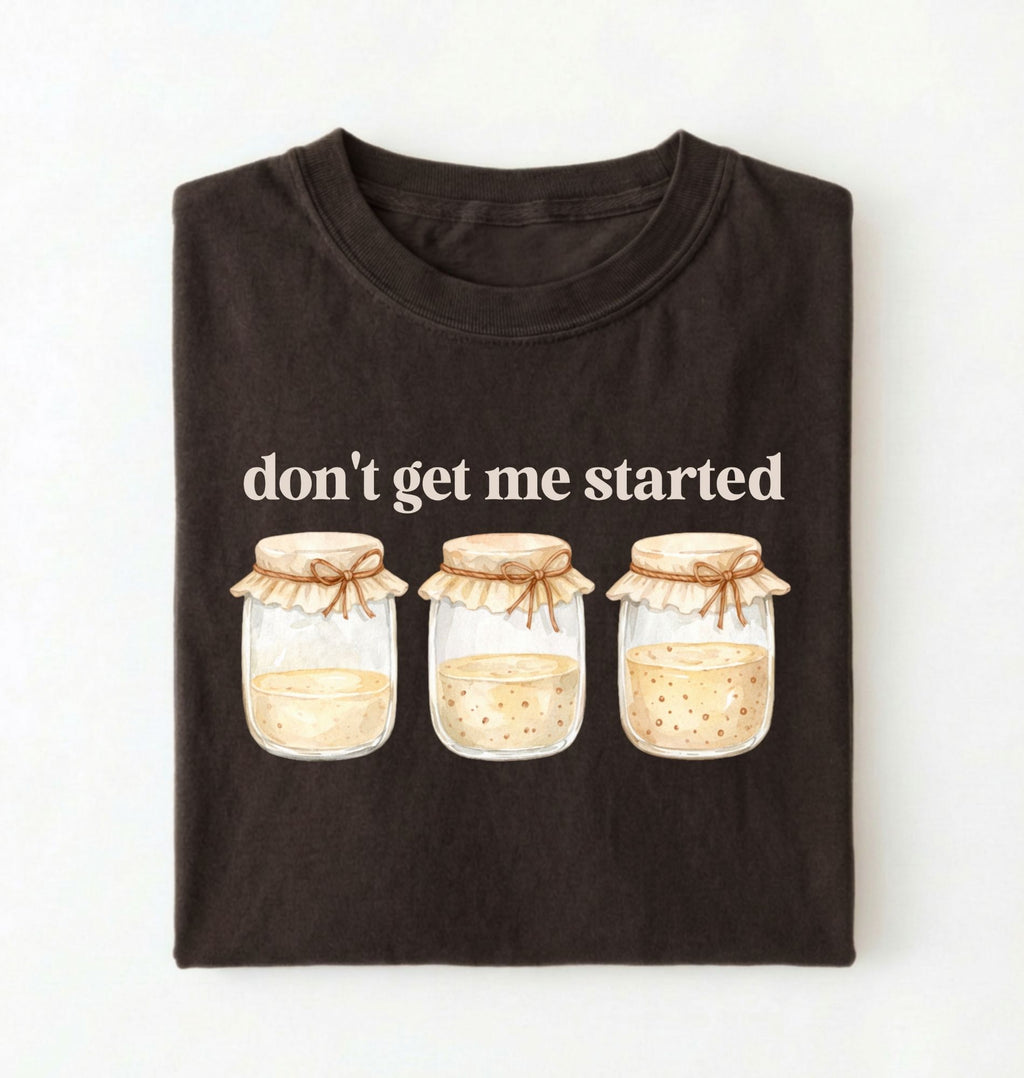 Flat lay of a dark chocolate t-shirt featuring illustrated jars and text reading “Don’t Get Me Started. Funny sourdough starter perfect gift for bakers fermentation lovers and bread enthusiasts on a white background
