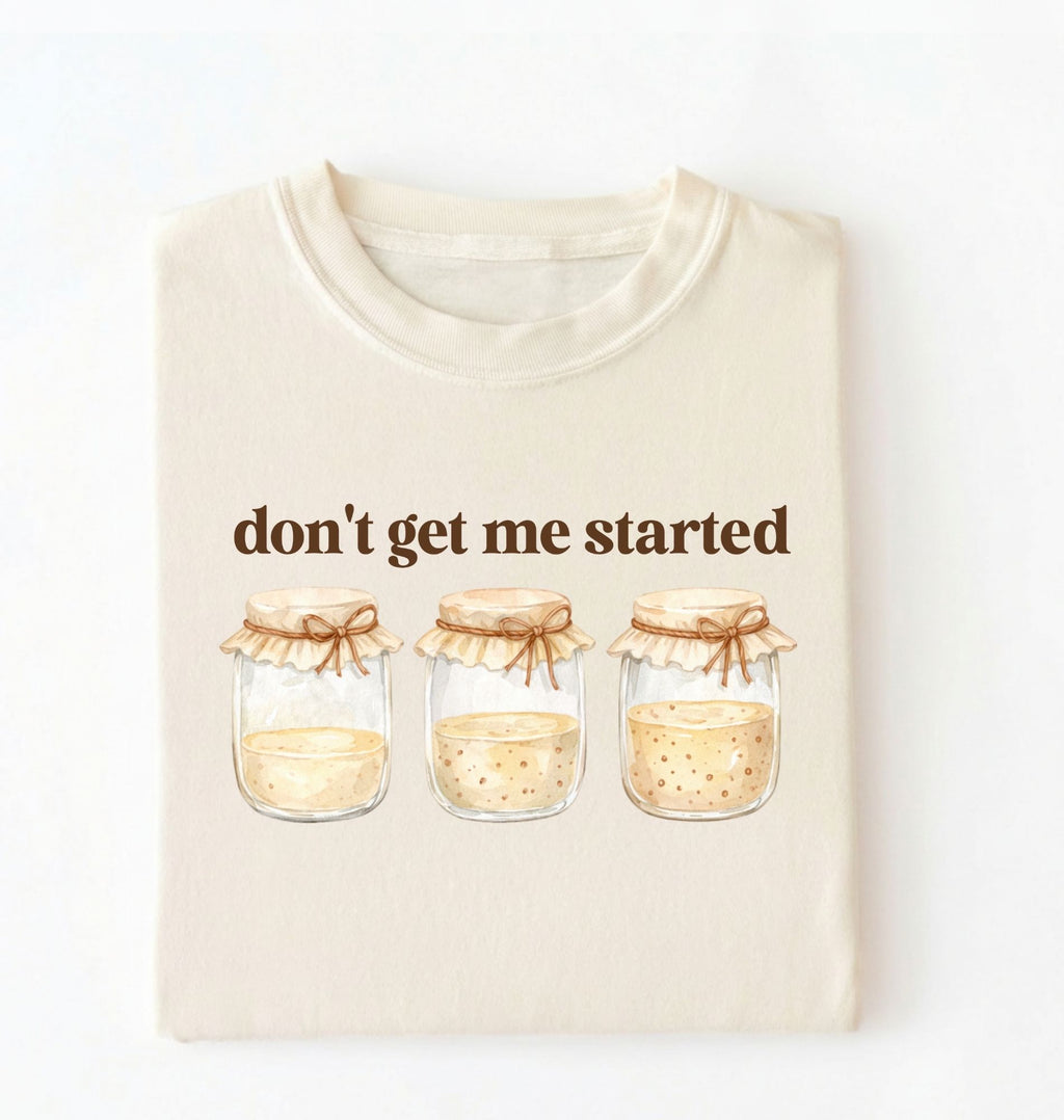 Flat lay of a ivory t-shirt featuring illustrated jars and text reading “Don’t Get Me Started. Funny sourdough starter perfect gift for bakers fermentation lovers and bread enthusiasts on a white background